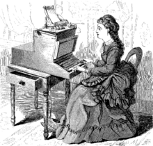 A woman in a long, frilled dress with a pleated train sits at a large, wooden typewriter which is approximately half her size. The typewriter is affixed to a small table with an open drawer.
