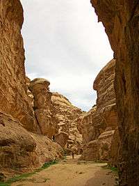 A narrow canyon with high, beige rounded stone walls and a dusty floor. A man can be seen walking in the distance