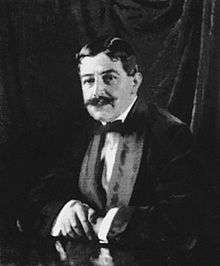 A Dark-haired gentleman with a bushy moustache leans, arms crossed on a table. He is wearing a velvet smoking jacket and bow-tie. A dark curtain is behind him.