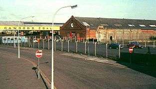 Fenced-off tarmac car parking area, with a run-down red-brick industrial building in the background.