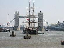 A sailing ship sits moored on the River Thames, with a large bridge in the background