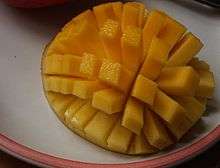 A halved, inside-out mango is cut in a grid pattern, still attached to the peel. The mango is inside-out, causing the resulting rectangles of fruit to splay out.