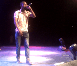 A picture of an African man on stage wearing short sleeve shirt, grey jeans, and boots. With his left hand he holds a microphone up to his mouth