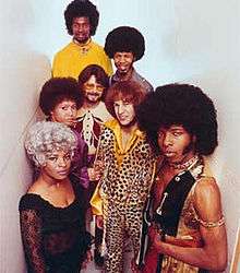 Seven young adults in garish clothes and hair. The most prominent is a black man in a vest with chains; he wears a large afro with sideburns, and looks with narrowed eyes and closed mouth at the camera. A black woman is in a platinum blonde wig and black dress. A white man with red hair wears a leopard print shirt and pants. There are two other black men, also in afros, another white man, with a short beard and glasses, and another black woman.