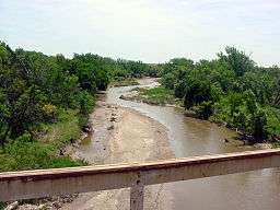 A small, muddy river, as seen from a bridge, meanders between tree-lined banks.