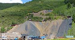 A wooded slope with two large bare patches of loose rock visible. At the bottom of the image are some vehicles and old temporary buildings. In the middle between the two are some wooden buildings and a flagpole with the American flag