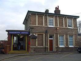 A red-bricked building with a rectangular, dark blue sign reading "SNARESBROOK STATION" in white letters all under a light blue sky