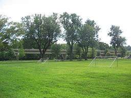 A green soccer field with trees and picnic tables along one edge and a highway bridge behind that