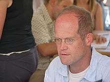 A color photograph of a seated, middle-aged man with a receding hairline. He is wearing spectacles, a white undershirt and a light blue collared shirt. He looks left past the camera.