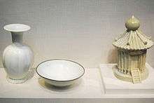Three pieces of white porcelain. From left to right, a vase with a grooved, fat body, a short, thin stem, and small opening with a large flat rim extending from it, a simple bowl with a thin brown trim around the outside rim, and a container in the shape of a granary, with several viable support posts, a tiled roof, and a staircase leading to a door, all built into the piece.