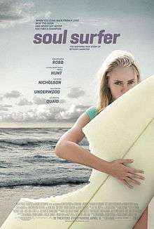 A young girl holds a surfboard at the beach. A section of her board is missing as if been bitten by a shark.
