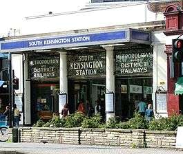 A white building with a rectangular, dark blue sign reading "SOUTH KENSINGTON STATION" in white letters all under a white sky