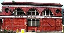 A two-storey red-glazed building with a single rectangular window on the lower floor and three large semi-circular windows on the upper floor flanked by two small circular windows. A white band between the floor levels displays "South Kensington station".