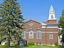 A side view of the same church. On a section with a pointed top and similar classical decoration are three large arched windows, the middle set with stained glass. A large evergreen tree blocks the view on the left