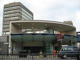 A grey building with a rectangular, dark blue sign reading "SOUTHWARK STATION" in white letters all under a light blue sky with white clouds