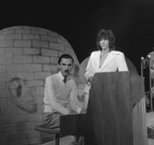 A black-and-white photo of two men standing at a piano, looking to the camera