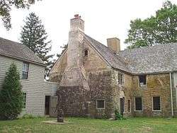 The rear of a 2+ story stone house has a wooden addition extending off to the left. An old-fashioned water pump with bucket is near the door.