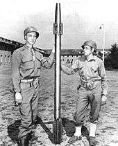 Two soldiers in US Army uniforms hold between them a long, slim projectile that is somewhat taller than them, with a finned tail
