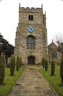 The tower of a stone church seen from the west, with a diagonal-shaped clock face, and an embattled parapet.
