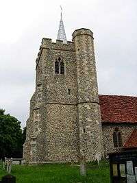 A flint tower with a battlemented parapet and a small spire; to the right is a stair turret rising higher than the tower