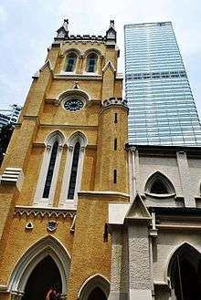 An ornate Gothic-style yellow church tower seen from below with a glass-surfaced skyscraper behind it.
