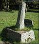 A very weathered square stone block with a short shaft leaning to the right