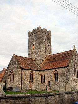Brown stone building with red roofs and central square tower. In the foreground are gravestones.