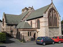 A church seen from the southeast, in light stone with darker bands; the tower is just visible; in front of the church are two parked cars