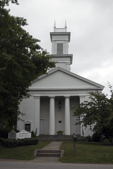 A white church with four narrow points atop its square steeple and columns in front of its entrance flanked by tall trees.