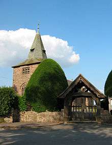 A tower with a broach spire partly obscured by bushes with a lych gate to the right and a wall and road in the foreground