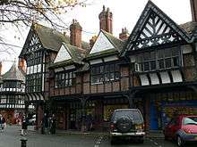 A row of shops seen at an angle with an arcade at ground level.  Above this the building is timber-framed, with four gables; the lateral two of these have painted plaster panels and decorated timber framing within the gable, and the central gables contain brick nogging and pargeting.