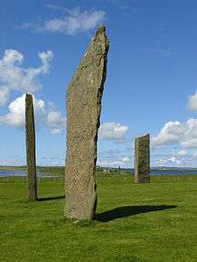 Three tall thin monoliths sit in a grassy field under blue skies with two bodies of water beyond at right and left.