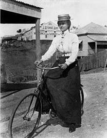 Woman, dressed in a long skirt and blouse with a hat, riding a bicycle along an unsealed road.