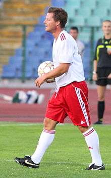 A photograph of a dark haired man on a football pitch with the football in his hands, the man is wearing a white shirt, red shorts and white socks.