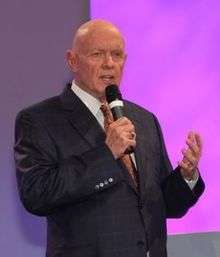 Older, bald man in a sharp suit speaking to an unseen audience