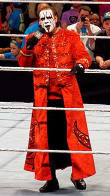 Sting wearing a red longcoat standing in a wrestling ring talking on a microphone