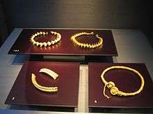 Three elaborate gold coloured torcs lie on dark red  surfaces. A fourth torc is in two pieces.