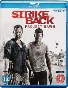 A blu-ray disc cover with a white/greying background and the title "Strike Back Project Dawn" in red letters on the top. Below it are two men. The man on the left is stubbly with a blue shirt and holding a machine gun in his right hand. The man on the right is wearing a dirty vest and holding a pistol in his left hand. Behind them is a city skyline with three helicopters flying overhead.