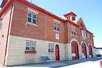 A two-storey concrete and red brick fire hall. The front of the hall has three large doors for vehicles, one regular door, and two windows.