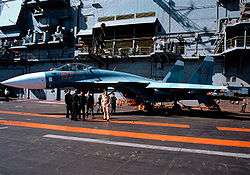 Aqua and blue jet aircraft on aircraft carrier deck, with a group of men standing close-by. Behind the jet is the ship's island
