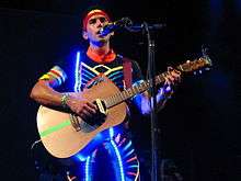 Stevens on-stage wearing a colorful suit and a string of lights while playing guitar and singing into a microphone