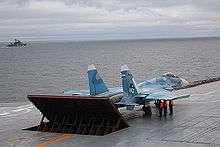 Rear port view of aqua-and-white jet aircraft lining up on aircraft carrier's deck, preparing for takeoff. The jet blast deflector is erected behind the aircraft. Three men in bright orange fluorescent tops stand underneath the jet's right wing