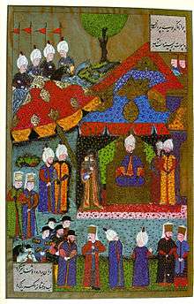A woman holding a baby in her arms stands before a man who wears a turban and sits on a throne in a tent which is surrounded by dozens of people