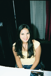 A black-haired woman wearing a yellow top and jeans sitting behind a table looking up at the camera.