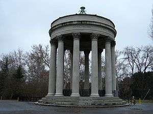 Open air round, marble structure resting on a layered base with twelve columns supporting the round, marble roof. There are leafless trees in the background.