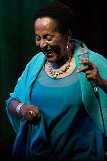 A woman in a blue dress holding a microphone with her right hand and smiling to the ground.