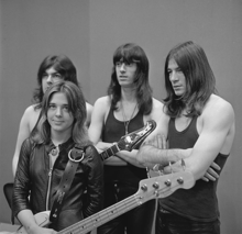 A black and white photograph of Quatro and her unnamed backing band. Quatro is holding her bass guitar, standing, and wearing a black leather jacket; her three taller and long-haired male bandmembers are standing behind her wearing dark tee shirts.