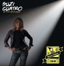 The front cover of Suzi Quatro's box set In the Spotlight Deluxe Edition. Quatro is standing up with her hands on her hips. A white spotlight shines on her from the top right, partly illuminating a black background. She is wearing a black leather jacket (over a black top) and light blue jeans with horizontal holes ripped in them. She has two black leather belts - a plain one, plus one decorated with yellow studs and yellow chains. In the bottom right  there is a yellow rectanglular logo with the words "Deluxe Edition" and "2 CD Set".