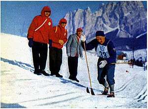 A man in a blue outfit with his competitor number "33" pinned to it skiing along a course in front of three other men.