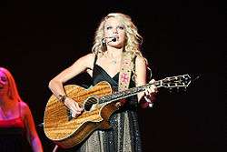 A female teen with blond hair and blue eyes, clothed by a sparkly silver dress, faces forward and plays a koa wood guitar.
